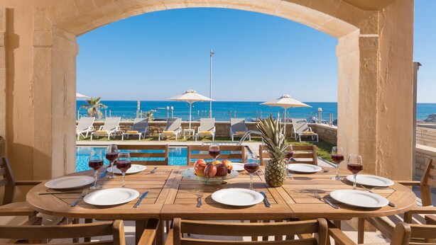 A dining table set for two with a view of the ocean and beach chairs in the background. - Irida beachfront Villa for 12 next to all amenities (Rethymno)