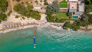 Oversigtsbillede af en kystejendom med en strand, swimmingpool og et stort hus med terrasse og udendørs møbler.