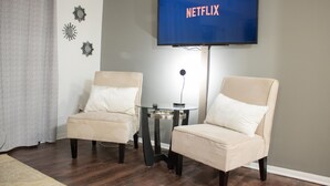 Un salon avec deux fauteuils beiges, une table basse en verre et un téléviseur à écran plat affichant le logo de Netflix.