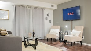 Un salon avec un canapé gris, deux fauteuils, une table basse en verre, un téléviseur à écran plat affichant le logo « NETFLIX » et un élément décoratif mural.