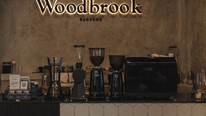 A coffee shop counter with a sign reading "Woodbrook Bangkok," various coffee-making equipment, and a tiled backsplash. - Hostel Urby (Bangkok)