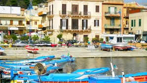 A coastal town with colorful boats docked at the marina, a prominent building with balconies, and a rocky cliff in the background.