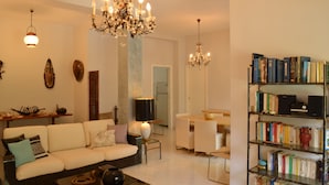 A modern living room with a sofa, a coffee table, a chandelier, a bookshelf, and a dining area.