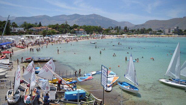 A crowded beach with clear turquoise water, boats, and a mountainous backdrop.