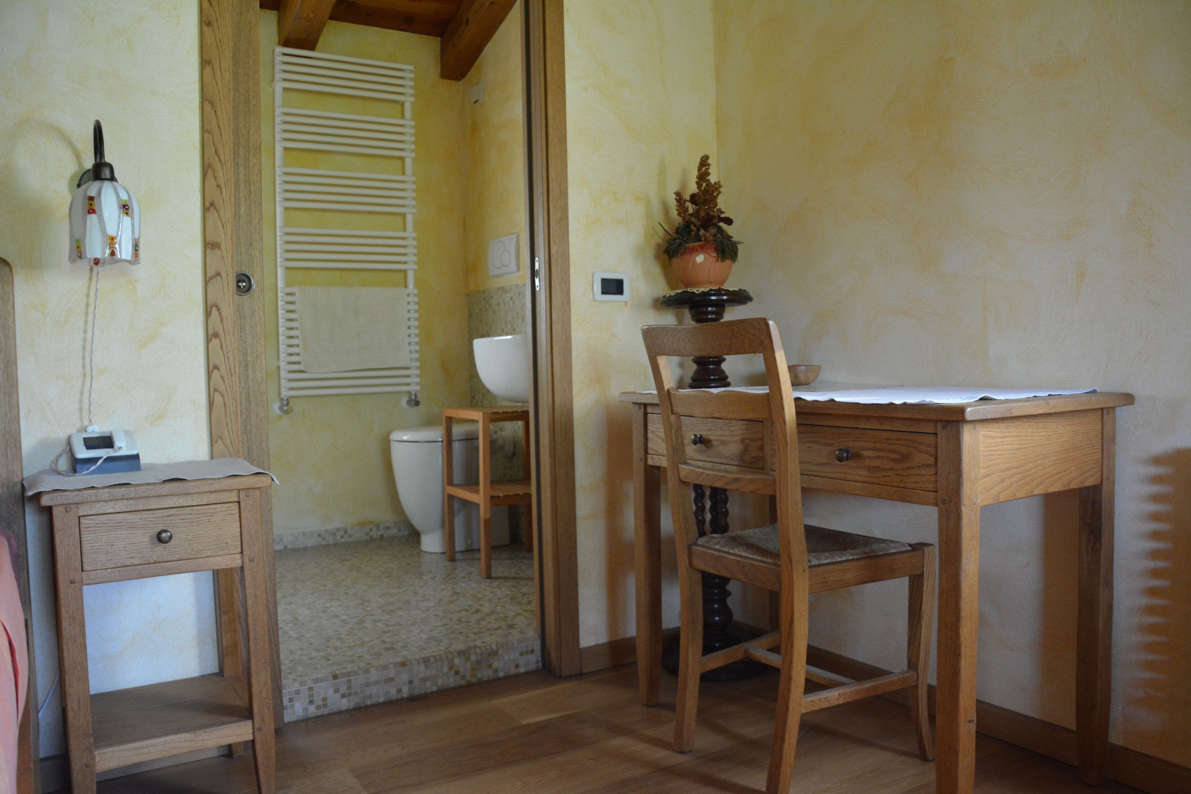 Quarto pequeno e compacto com escrivaninha de madeira, cadeira, mesa de cabeceira e sanitário.