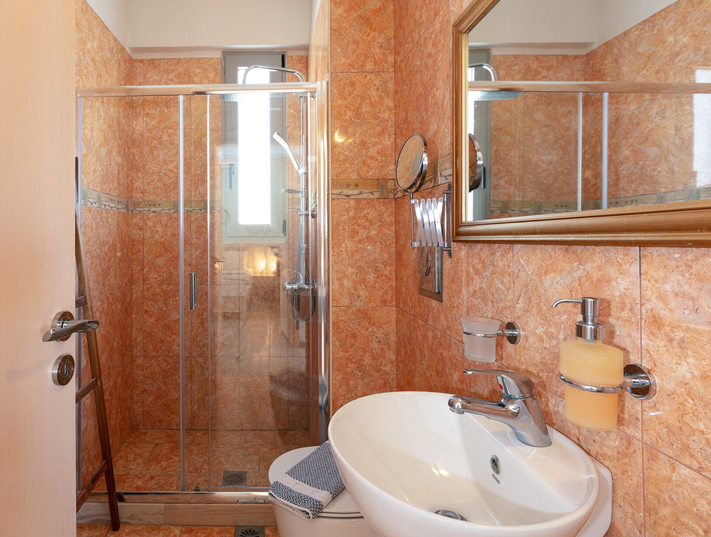 Un baño con ducha envuelta en vidrio, un lavabo blanco con accesorios cromados y un espejo grande sobre el lavabo.