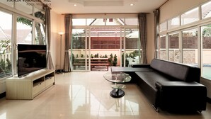 A modern living room with a flat-screen TV, a glass coffee table, a black sofa, and large windows offering a view of the outside. - Hansa Paradise Hill Pool Villa (Pattaya)