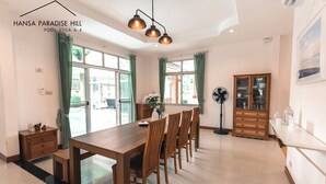 A spacious dining area with a long wooden table and chairs, a modern ceiling light fixture, and a view of the outdoors through large windows. - Hansa Paradise Hill Pool Villa (Pattaya)