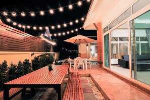 An outdoor patio with wooden tables and chairs, string lights, and a glass wall. - Hansa Paradise Hill Pool Villa (Pattaya)
