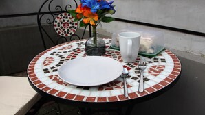 A round table with a mosaic tablecloth, a white plate, a fork, a spoon, a clear mug, and a vase with flowers.