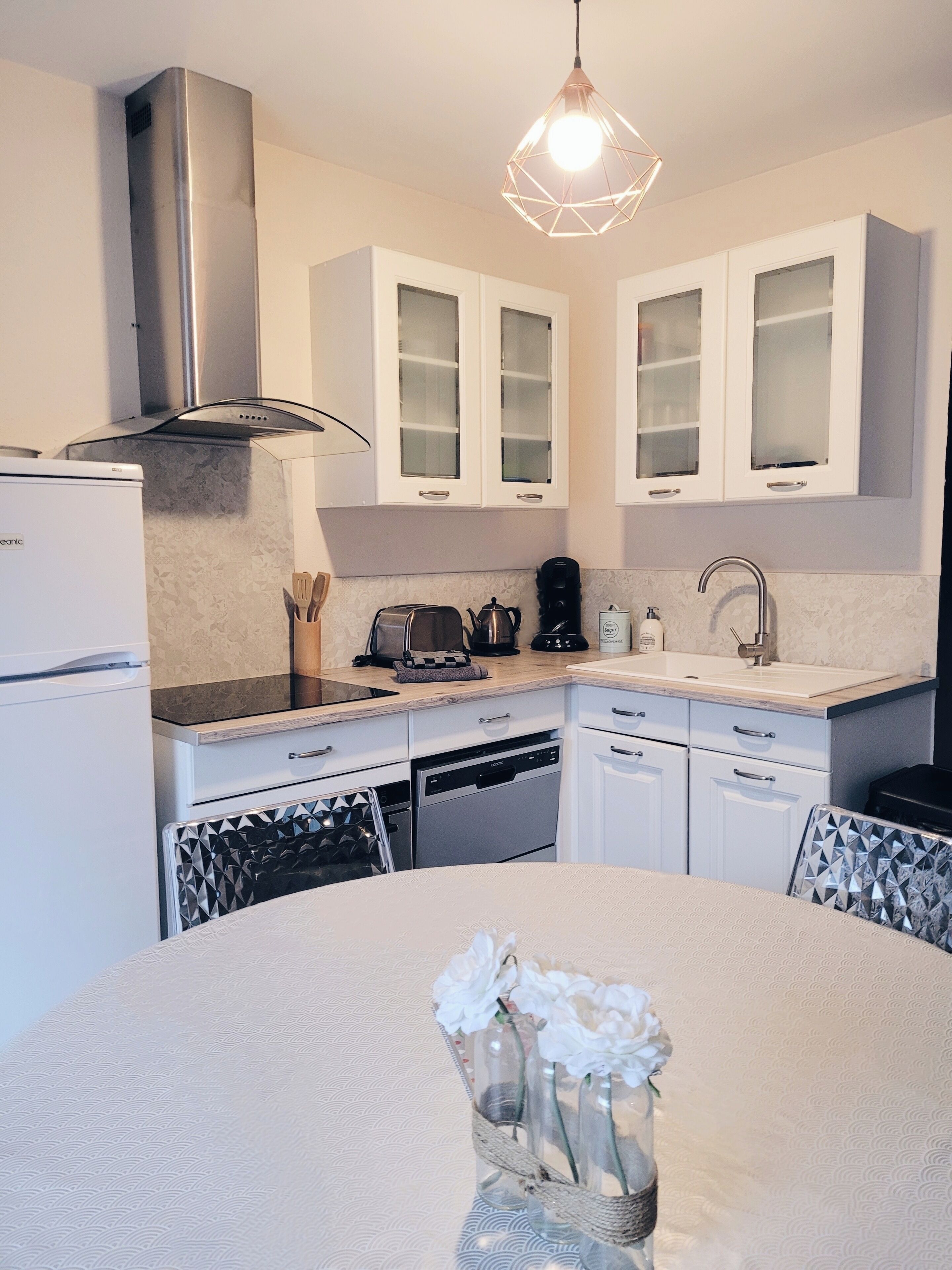 A modern kitchen with white cabinetry, a stainless steel range hood, a built-in dishwasher, and a dining area with a round table and a vase of flowers.