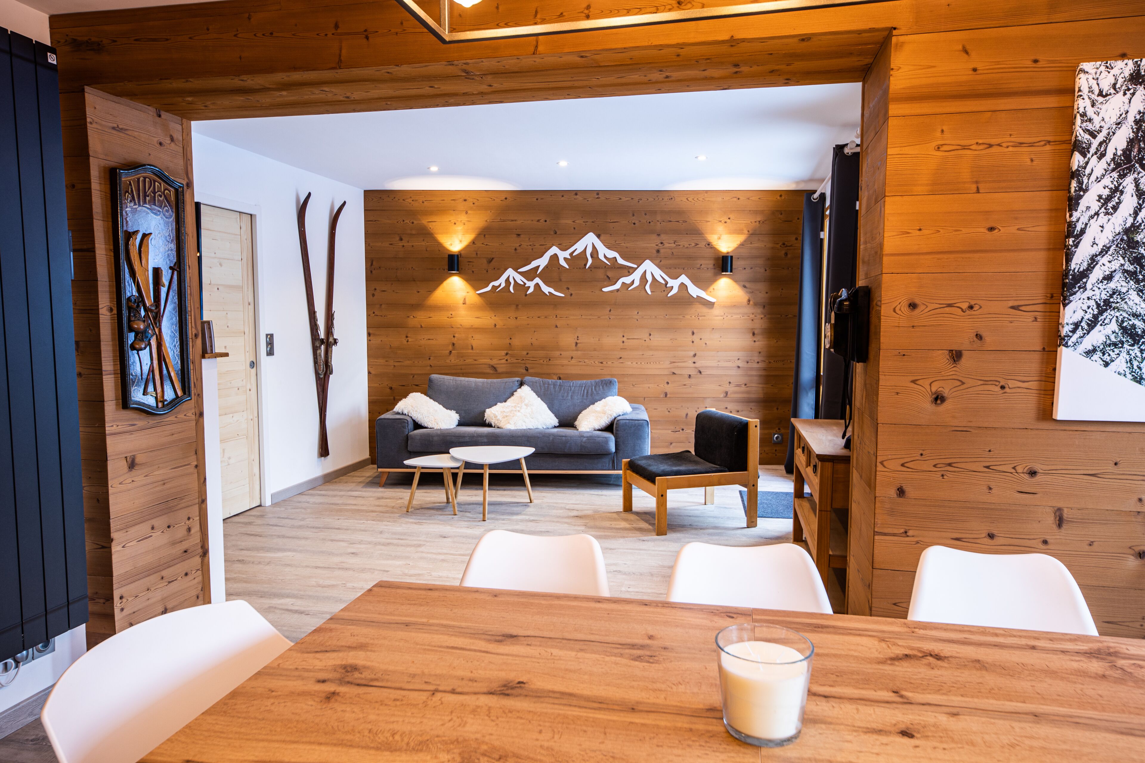 A cozy room with a wooden ceiling, a grey sofa with white cushions, a wooden dining table with chairs, and a glass of milk.