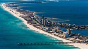 An aerial view of a coastal resort area with a long sandy beach, a pier extending into the ocean, and a cluster of buildings along the shore. - Gulf Front Condo with Luxury Furnishings and Beach Chairs  (Pensacola Beach)