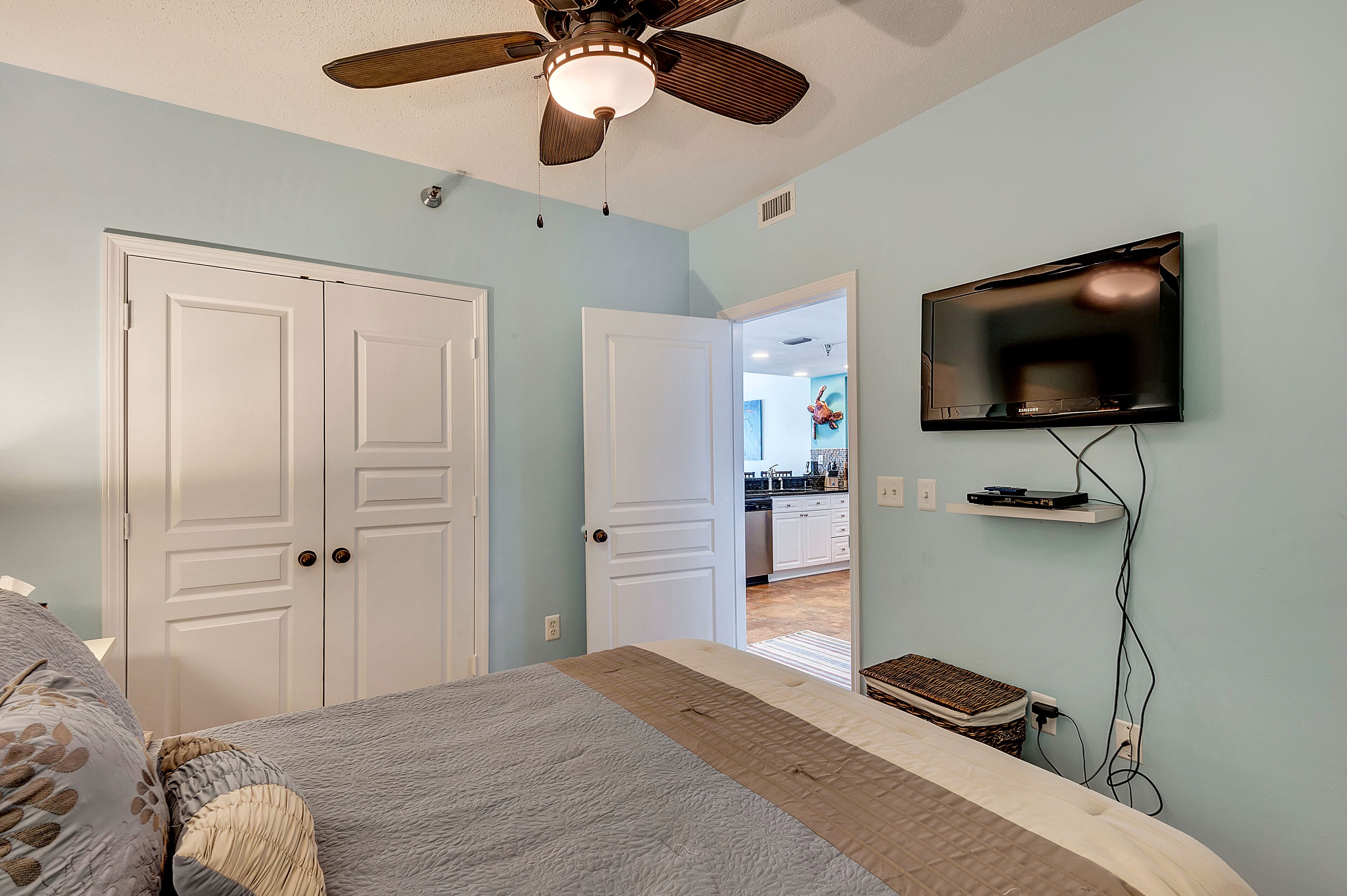 A bedroom with a bed, a ceiling fan, a TV mounted on the wall, and a door leading to a kitchen.
