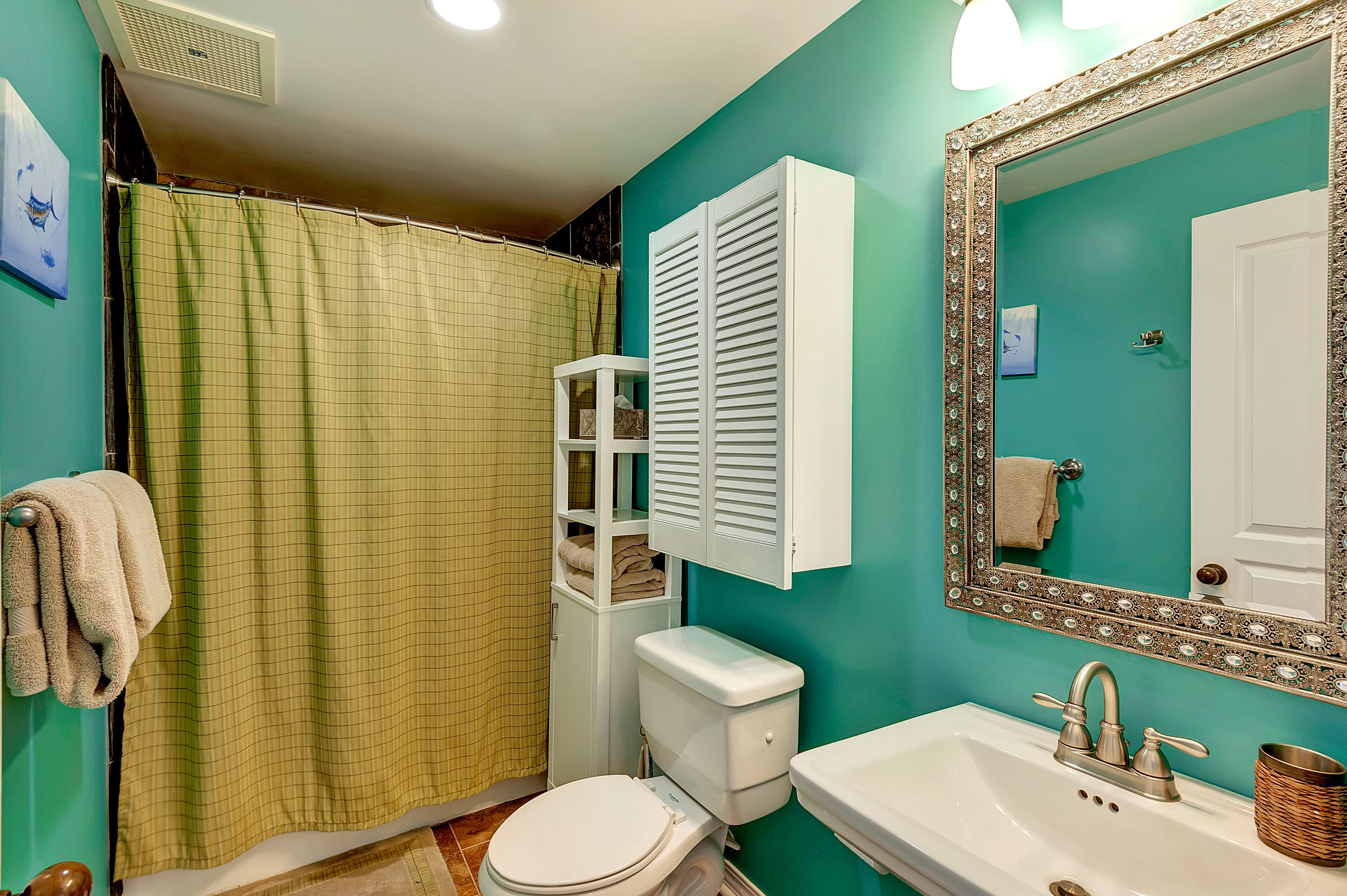 Un baño con pared verde, inodoro blanco, lavamanos con espejo y cortina de ducha.