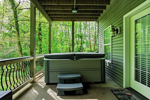 A covered porch with a hot tub, a wooden bench, and a railing overlooking a forested area.
