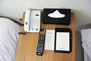 A hotel room with a wooden nightstand holding a telephone, a remote control, a no-smoking sign, and a black folder.