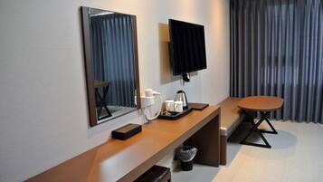 A hotel room with a wooden desk, a flat-screen TV, a mirror, a bench, and a small table.