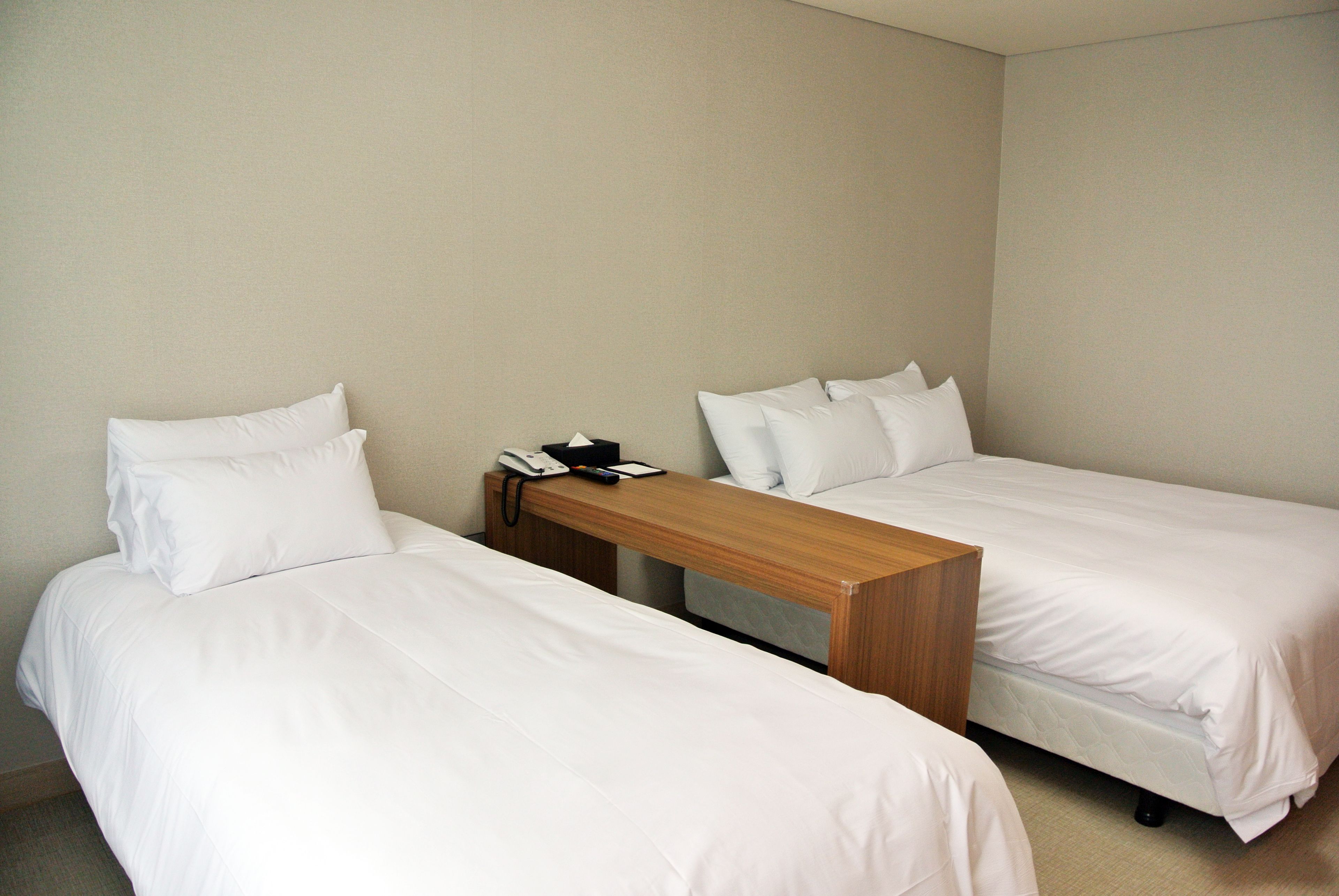 A hotel room with two single beds, a wooden nightstand with a telephone and notepad, and a plain wall.