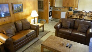 A living room with two brown leather sofas, a wooden coffee table, and a lamp. There is a kitchen area with wooden cabinets and a microwave.