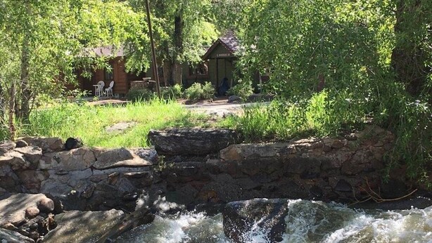 A rustic stone bridge over a flowing river, surrounded by lush greenery and a small wooden cabin in the background.