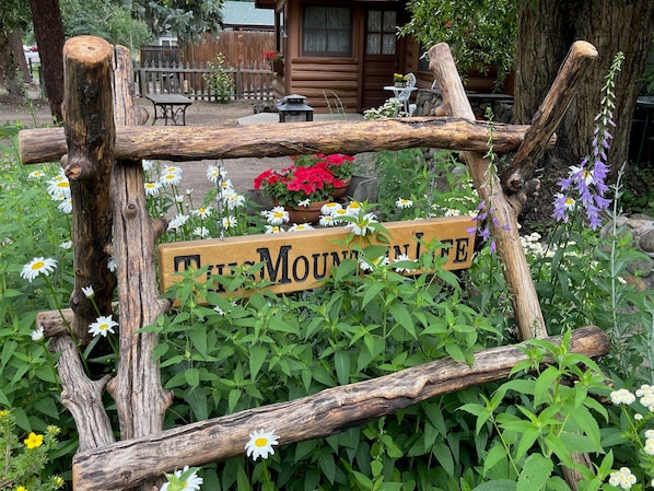 Ein rustikales Holzschild mit dem Schriftzug 'THE MOUNTAIN LIFE' inmitten eines Gartens mit verschiedenen Blumen und Grünpflanzen.