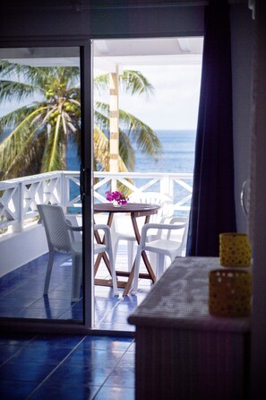 A room with a view of the ocean, a table with chairs, and a small table with a vase of flowers.