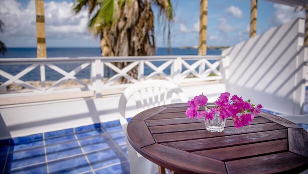 Una mesa de madera con un jarrón de vidrio con flores rosadas en un balcón con vistas a una piscina y al océano.
