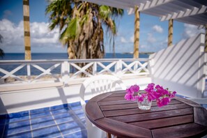 Een houten tafel met een glazen vaas vol roze bloemen op een balkon met uitzicht op een zwembad en de oceaan.