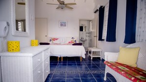 Un dormitorio con una cama, un tocador blanco, un banco, un ventilador de techo y un baño visible a través de una puerta abierta.