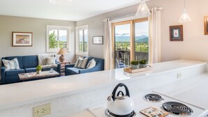 Eine moderne Küche mit weißem Arbeitsplatz, einem Wasserkocher und einem Herd. Ein blaues Sofa bietet Blick auf die Berge durch das Fenster.