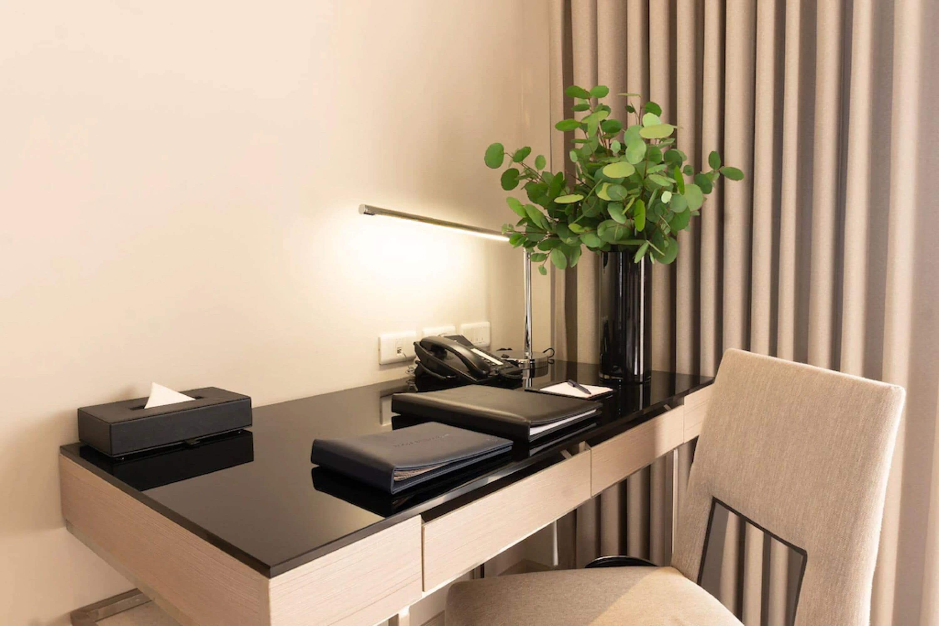 A modern hotel desk with a black surface, a vase with green leaves, a black telephone, and a beige chair.