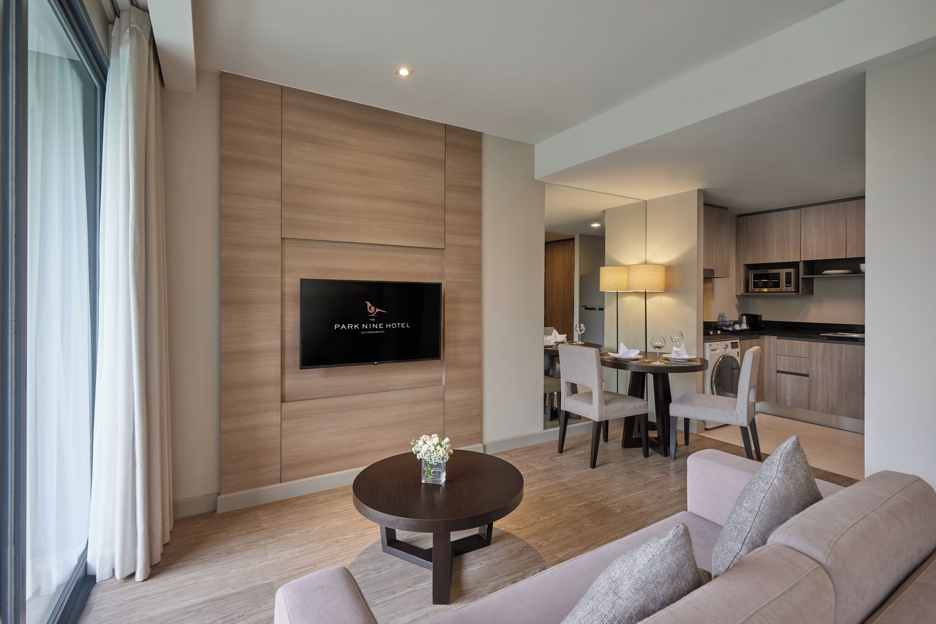 Una habitación de hotel moderna con televisor de pantalla plana montado en la pared, una mesa redonda con sillas, un sofá y una cocina con microondas y armarios.