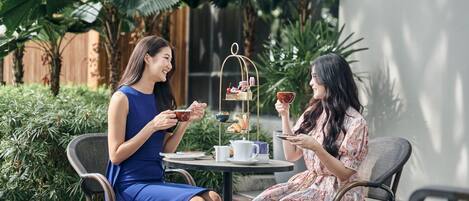 Two women seated at an outdoor table with a tea service, surrounded by lush greenery.