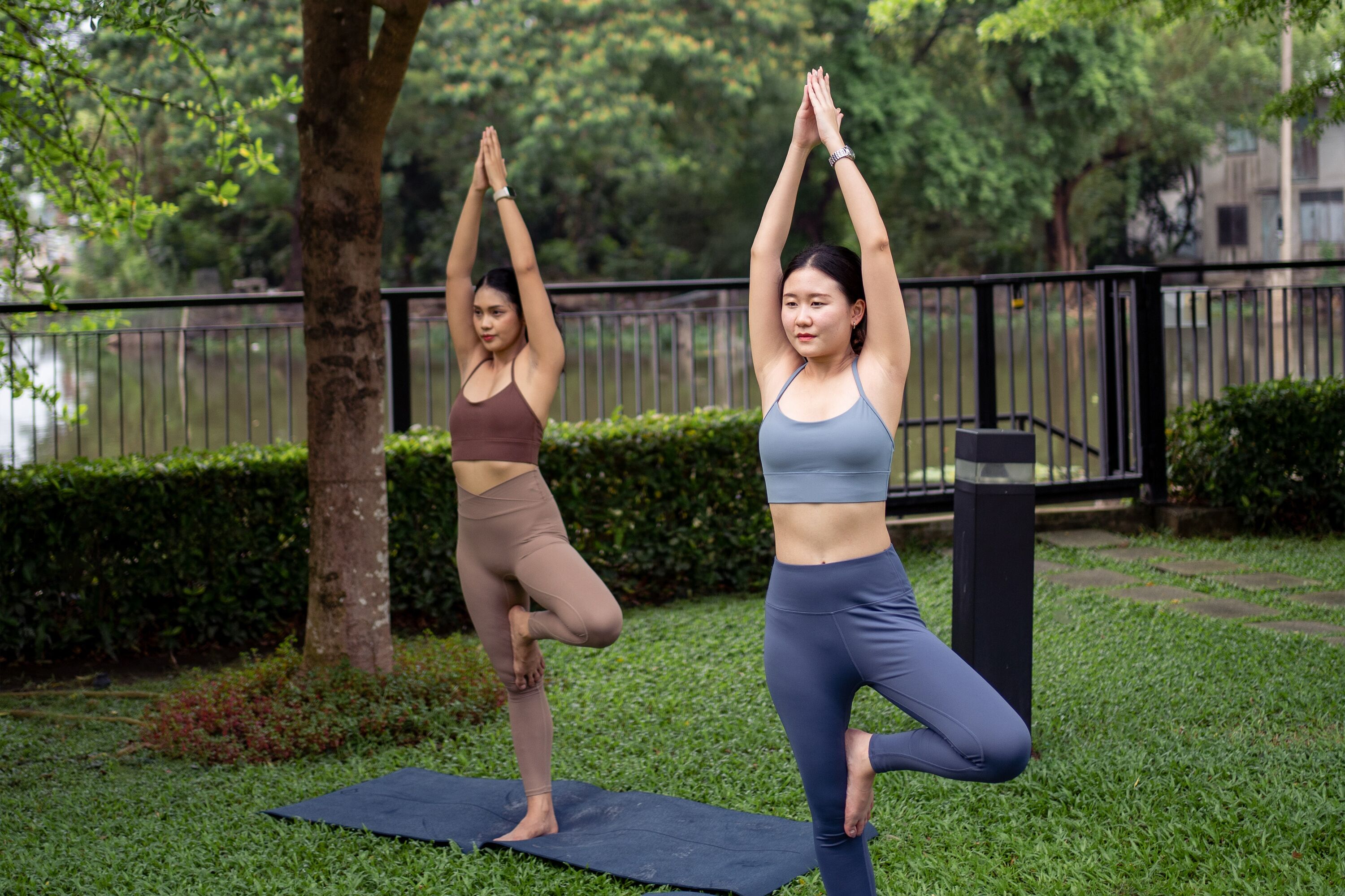 Dos personas practicando yoga sobre una esterilla en un parque, con una cerca y vegetación al fondo.