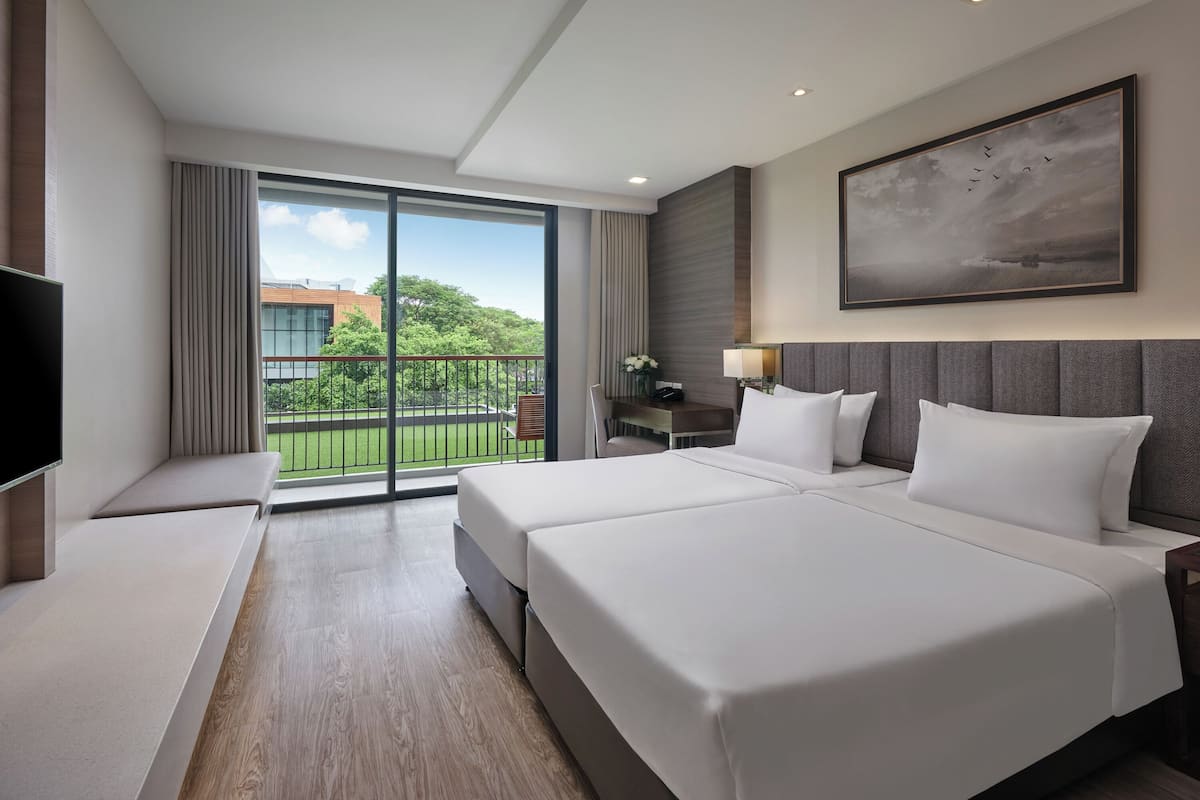 Ein Hotelzimmer mit einem großen Bett, einem Fernseher, einem Schreibtisch und einem Balkon mit Blick auf Grünflächen und Gebäude.