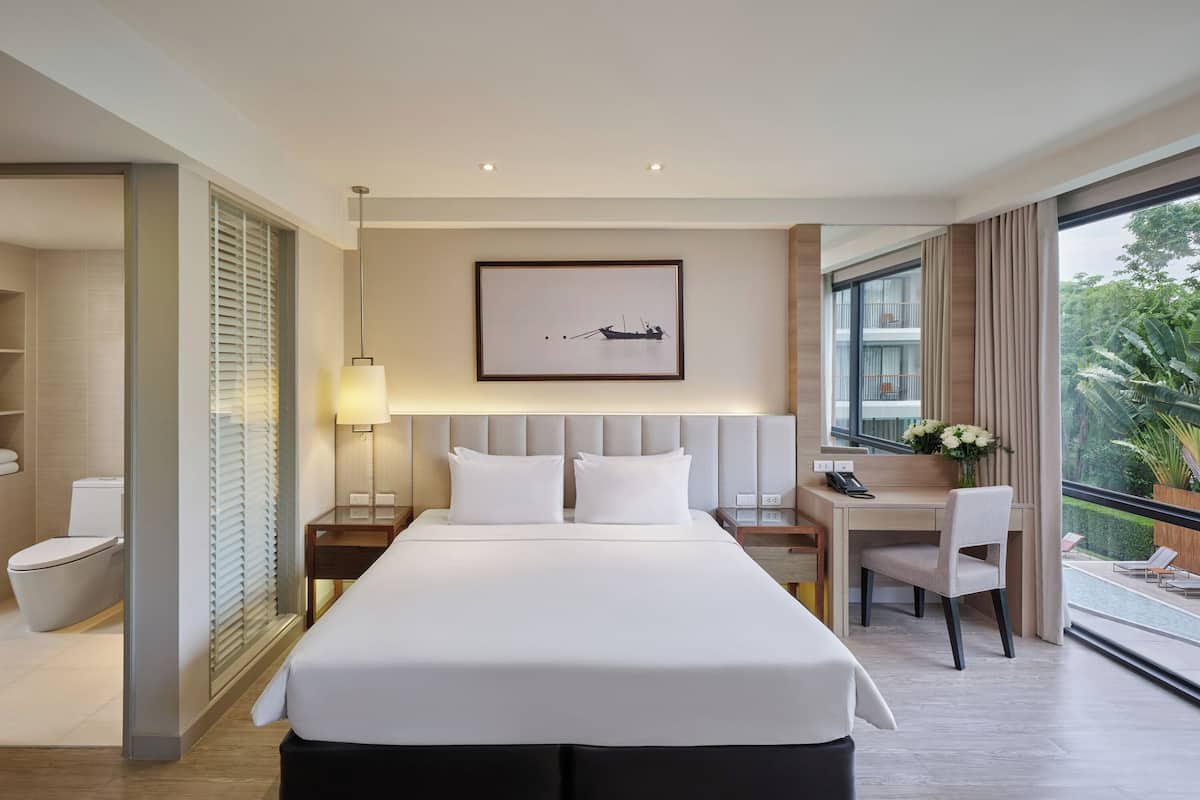 Ein modernes Hotelzimmer mit einem großen Bett, einem Schreibtisch mit Stuhl, einem Nachttisch mit Lampe und Blick ins Freie.