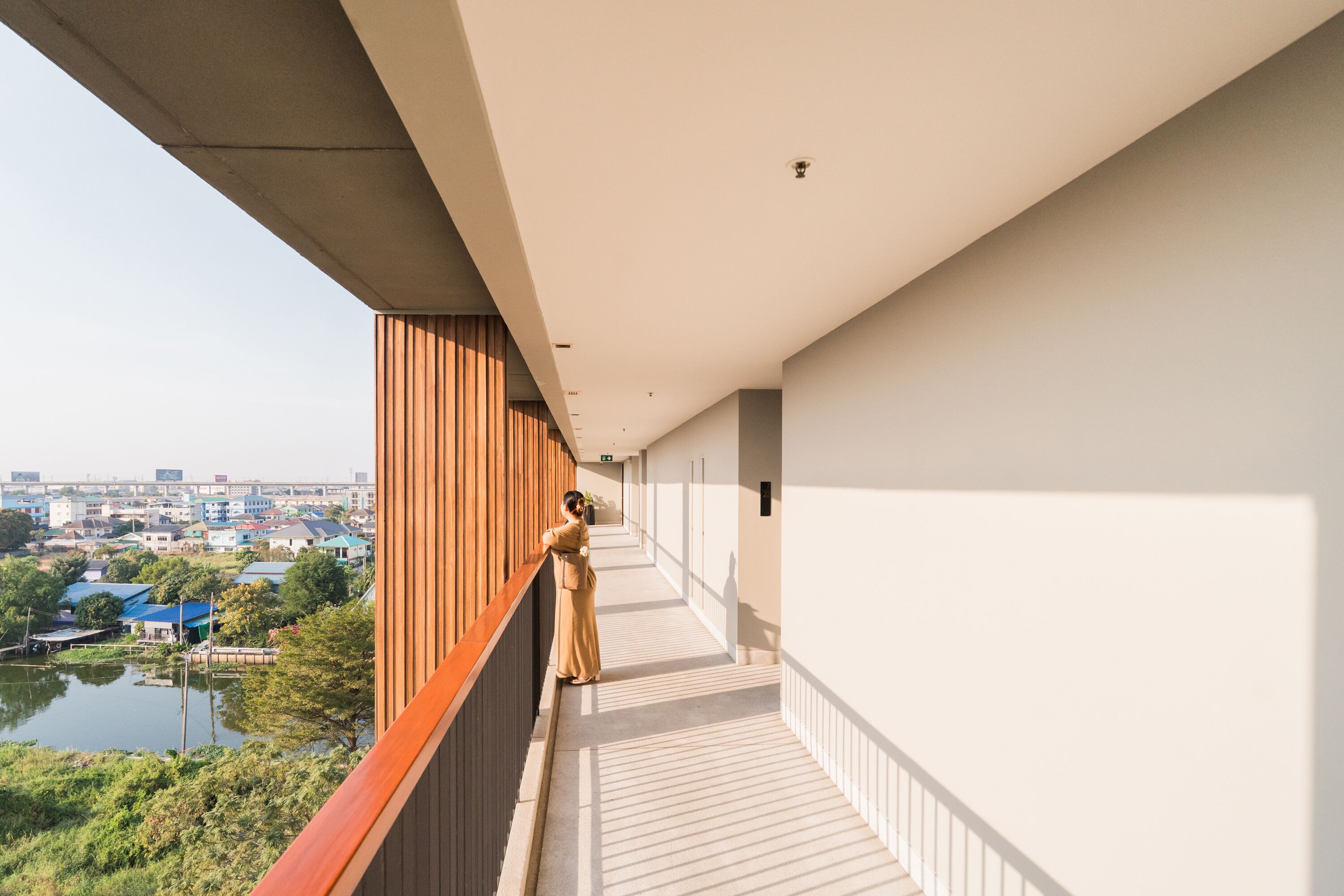 A corridor with a wooden railing on one side and a white wall on the other, leading to a balcony with a view of a body of water and buildings.