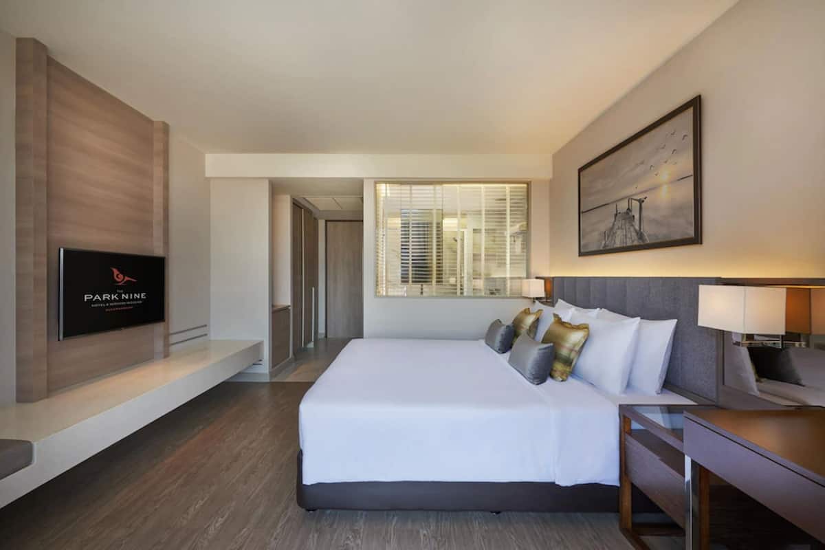 Ein modernes Hotelzimmer mit einem großen Bett, einem an der Wand montierten Fernseher, einem Schreibtisch mit Stuhl und einem Fenster mit Jalousien.