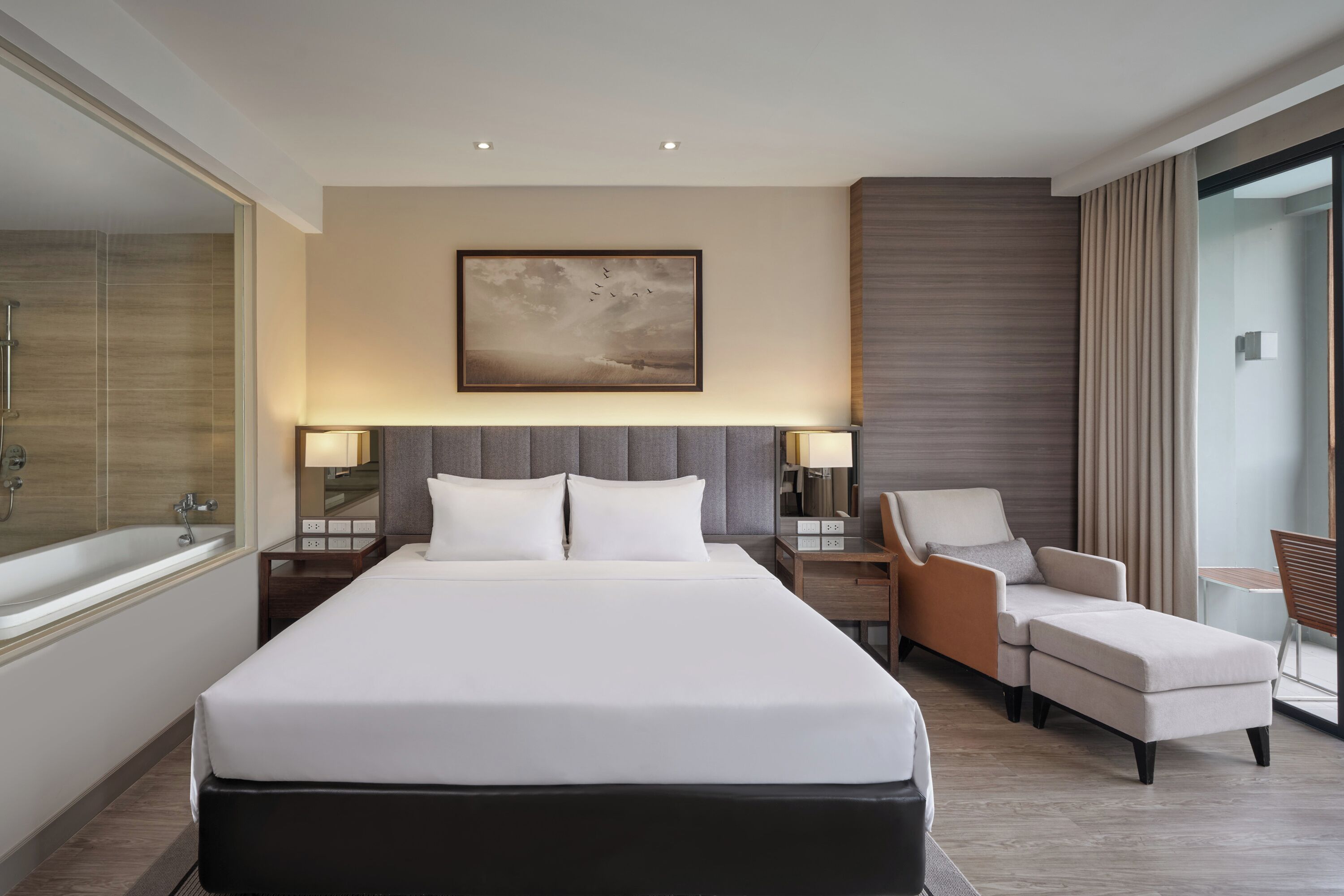 A modern hotel room with a large bed, a sitting area with a chair, a small table, and a large window offering a view.