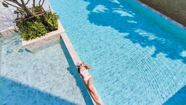 A woman lying on the edge of a swimming pool with clear water, surrounded by greenery and a stone structure. - The Park Nine Hotel Suvarnabhumi  (Bangkok)