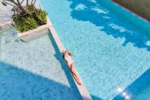Une femme allongée au bord d’une piscine à l’eau claire, entourée de verdure et d’une structure en pierre.