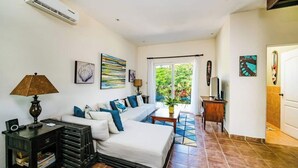 A modern living room with a sofa, a coffee table, and a TV. There is a window with a view of greenery outside. - La Cometa 19♆ Private rooftop terrace convenient in ♥ of town (Tamarindo)
