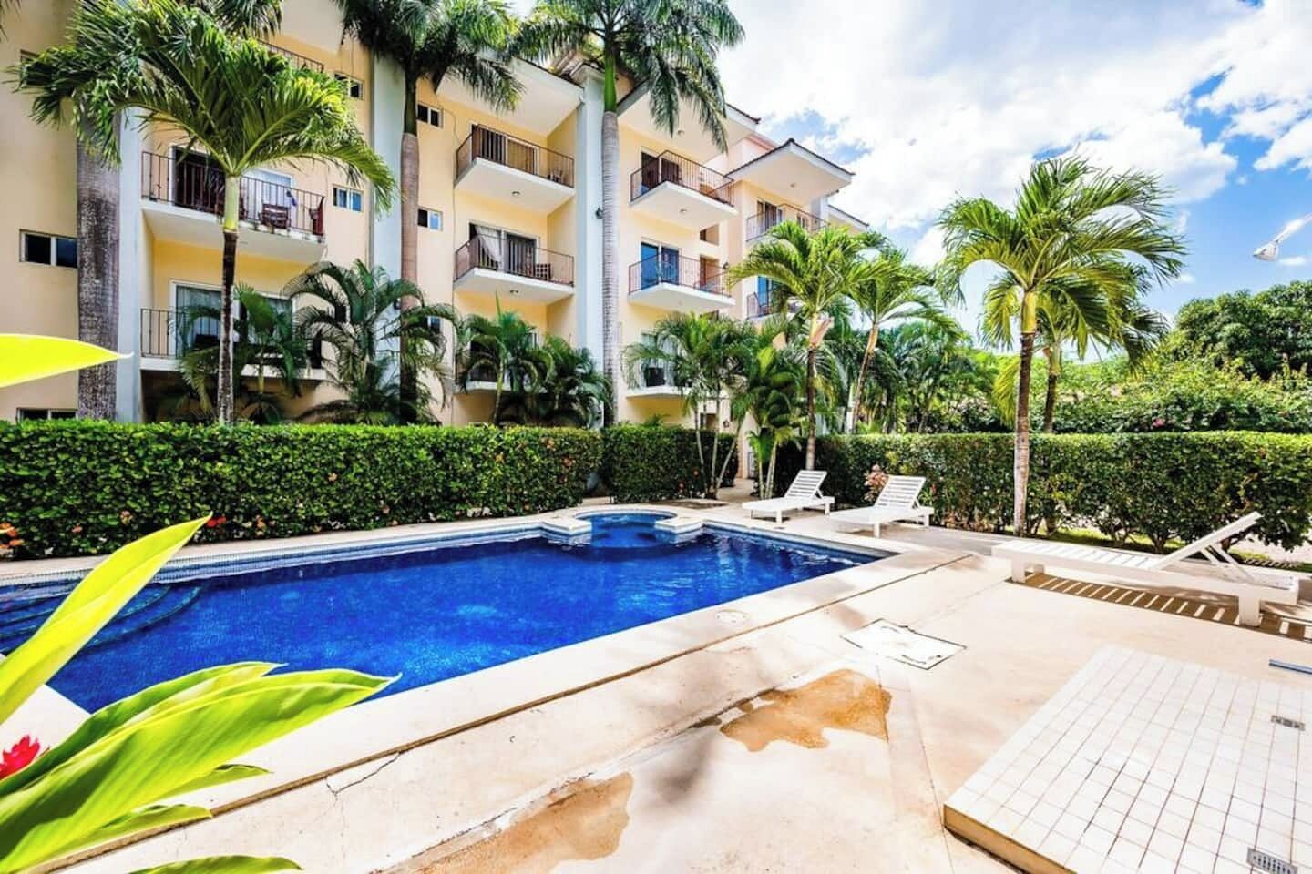 A swimming pool surrounded by palm trees and a multi-story building with balconies.