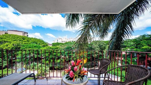 A balcony with wicker chairs, a small table with a flower arrangement, and a view of lush greenery and buildings in the background. - La Cometa 19♆ Private rooftop terrace convenient in ♥ of town (Tamarindo)