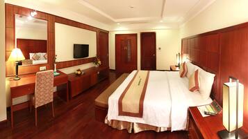 A hotel room with a large bed, a desk with a lamp, a television, and a wardrobe.