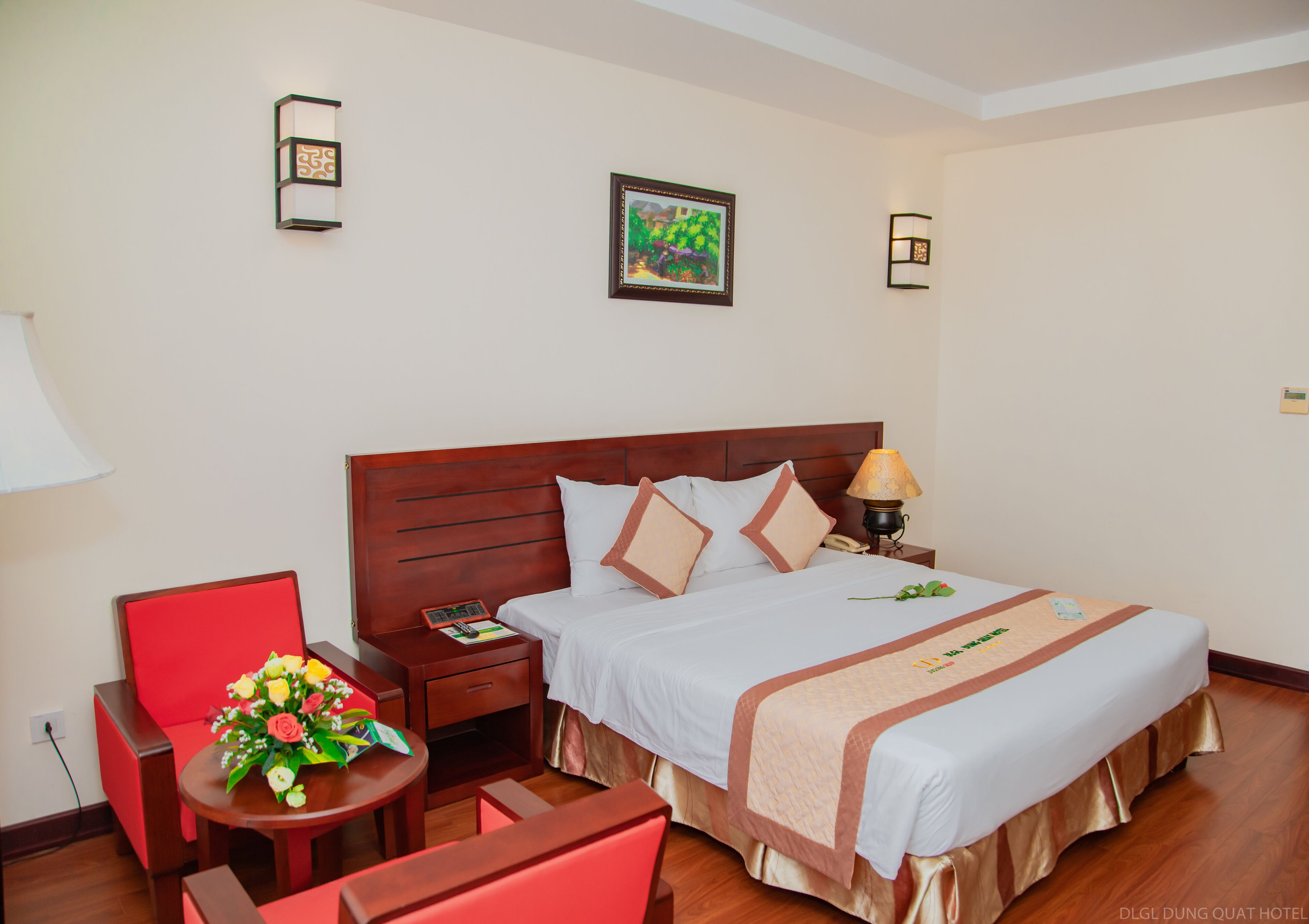 A hotel room with a wooden bed, a red chair, a small table with a flower arrangement, and a nightstand with a lamp.