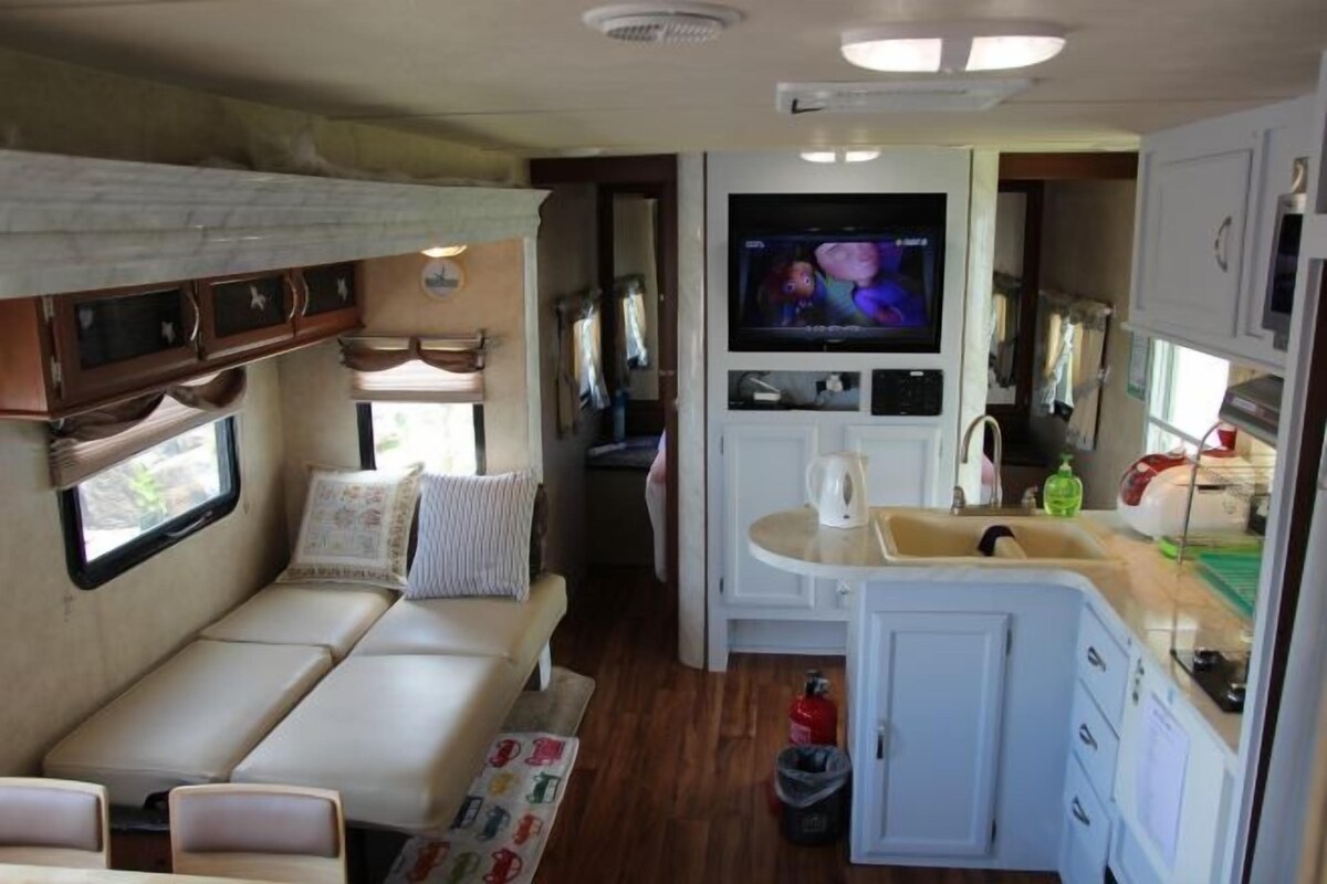 A compact RV interior with a sofa, a television, a kitchenette, and a small dining area.