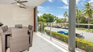 A balcony with wicker furniture, a ceiling fan, and a view of a parking lot with cars and palm trees. - Peponi 4 - Beachfront Apartment (Yorkeys Knob)