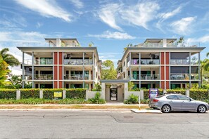 Un edificio de apartamentos moderno con varios balcones, entrada protegida por un portón y un coche estacionado.